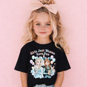 A young girl with blonde hair wearing a black t-shirt with a graphic of two cartoon girls and the text "Girls Just Wanna Have Fun" is posing against a pink background.