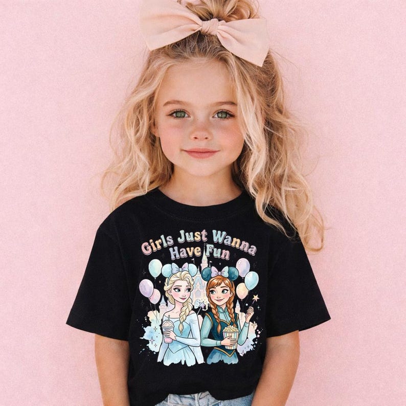 A young girl with blonde hair wearing a black t-shirt with a graphic of two cartoon girls and the text "Girls Just Wanna Have Fun" is posing against a pink background.