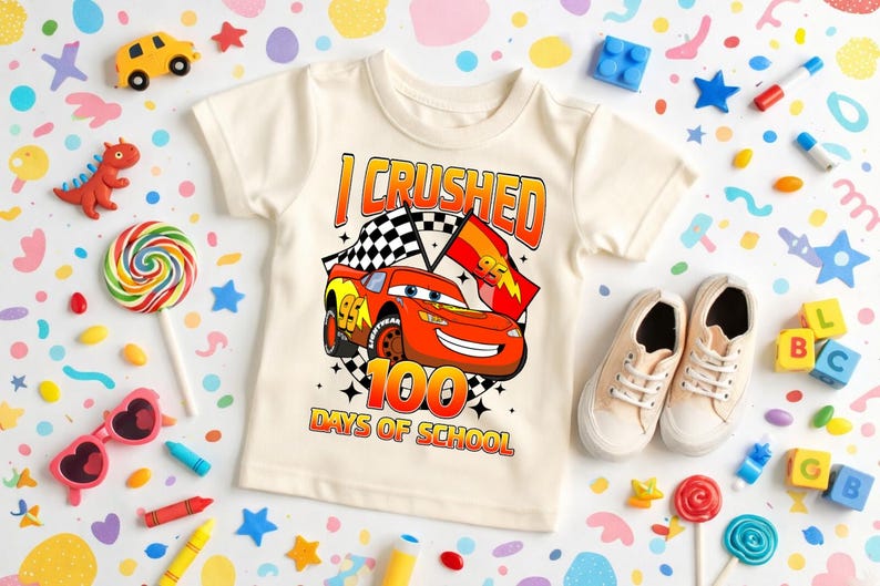 a white t-shirt with a graphic of a race car and the text "I CRUSHED 100 DAYS OF SCHOOL", surrounded by various children's toys and items.