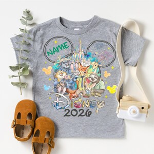 A gray t-shirt with a Disney-themed graphic, a pair of brown baby shoes, and a camera are arranged on a white background.