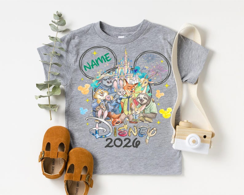 A gray t-shirt with a Disney-themed graphic, a pair of brown baby shoes, and a camera are arranged on a white background.