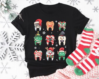 May include: Black t-shirt with a Christmas-themed design featuring cartoon teeth wearing festive hats and accessories. The teeth are decorated with candy canes, bows, and other holiday elements.