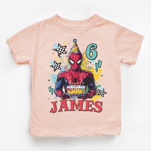 A pink t-shirt with a graphic design featuring a Spider-Man character holding a birthday cake, surrounded by stars and lightning bolts.