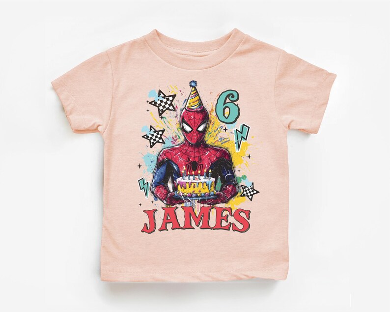 A pink t-shirt with a graphic design featuring a Spider-Man character holding a birthday cake, surrounded by stars and lightning bolts.