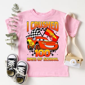 A pink t-shirt with a graphic of a race car and the text "I CRUSHED 100 DAYS OF SCHOOL" printed on it, along with a pair of black and white sneakers and a camera.