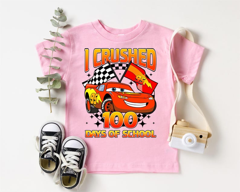 A pink t-shirt with a graphic of a race car and the text "I CRUSHED 100 DAYS OF SCHOOL" printed on it, along with a pair of black and white sneakers and a camera.