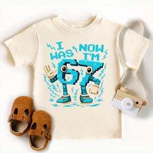 A white t-shirt with a graphic design, a pair of brown baby shoes, and a white camera are arranged on a white background.
