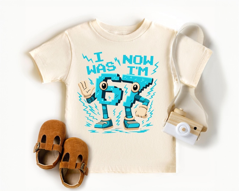 A white t-shirt with a graphic design, a pair of brown baby shoes, and a white camera are arranged on a white background.