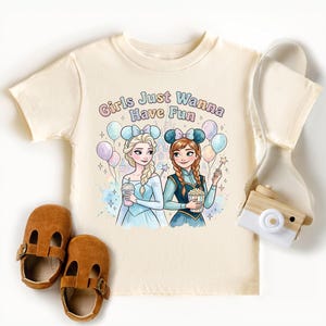 A white t-shirt with a graphic design featuring two cartoon girls, one blonde and one brunette, surrounded by balloons and holding drinks. The text on the t-shirt reads "Girls Just Wanna Have Fun".
