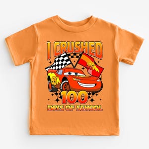 An orange t-shirt with a graphic design featuring a cartoon car and the text "I CRushed 100 DAYS OF SCHOOL".