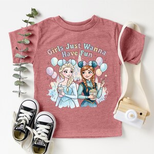 A pink t-shirt with a graphic of two cartoon girls, one blonde and one brunette, surrounded by balloons and holding drinks, along with the text "Girls Just Wanna Have Fun". The t-shirt is displayed on a white background, with a pair of black and white sneakers, a white camera strap, and some greenery.