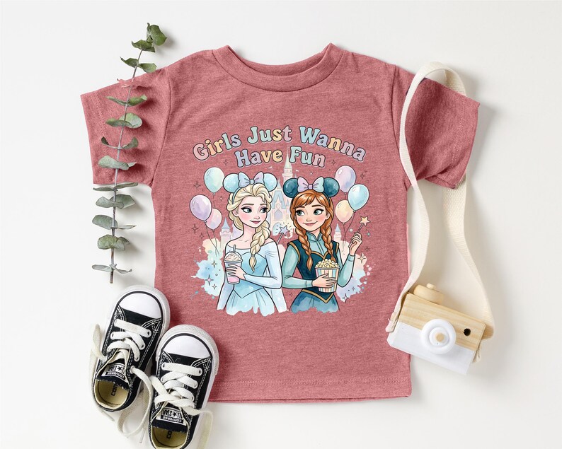 A pink t-shirt with a graphic of two cartoon girls, one blonde and one brunette, surrounded by balloons and holding drinks, along with the text "Girls Just Wanna Have Fun". The t-shirt is displayed on a white background, with a pair of black and white sneakers, a white camera strap, and some greenery.