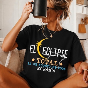 Può includere: Una t-shirt nera con un design grafico che presenta una mezzaluna e una stella gialle, la scritta bianca EL ECLIPSE, la scritta arancione TOTAL e la scritta azzurra 12 DE AGOSTO DE 2026 ESPAÑA, con il contorno bianco della Spagna.