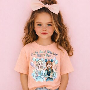 A young girl with long brown hair wearing a pink bow and a pink t-shirt with a graphic of two cartoon girls.
