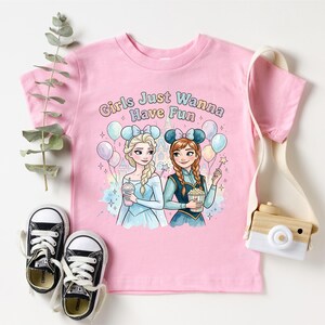A pink t-shirt with a cartoon illustration of two girls, one blonde and one brunette, surrounded by balloons and holding drinks, along with the text "Girls Just Wanna Have Fun". The t-shirt is placed on a white background, and there are a pair of black and white sneakers, a camera, and a plant nearby.