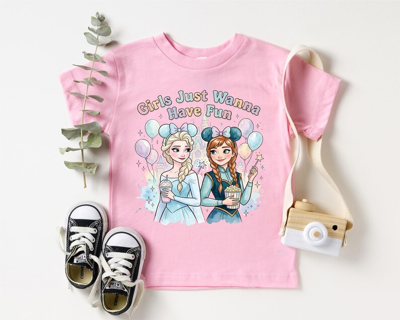 A pink t-shirt with a cartoon illustration of two girls, one blonde and one brunette, surrounded by balloons and holding drinks, along with the text "Girls Just Wanna Have Fun". The t-shirt is placed on a white background, and there are a pair of black and white sneakers, a camera, and a plant nearby.