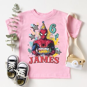 A pink t-shirt with a Spiderman graphic and the number 6, along with a pair of sneakers and a camera, all arranged on a white background.