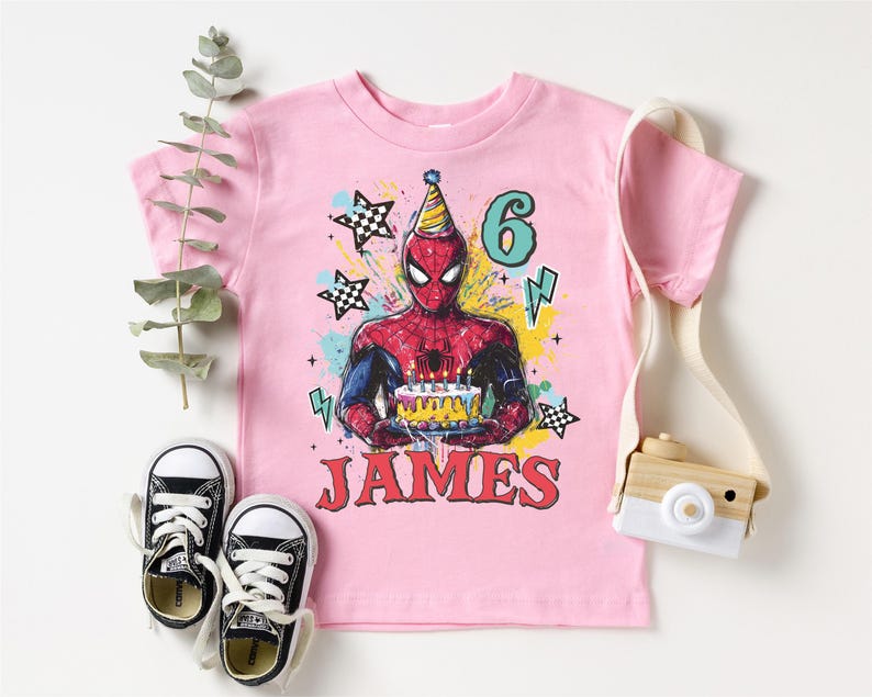 A pink t-shirt with a Spiderman graphic and the number 6, along with a pair of sneakers and a camera, all arranged on a white background.