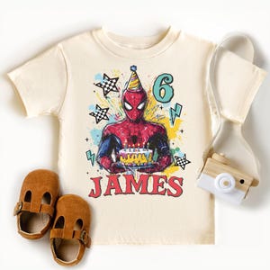 A white t-shirt with a Spiderman graphic and the number 6, along with brown baby shoes and a camera, all arranged on a white background.