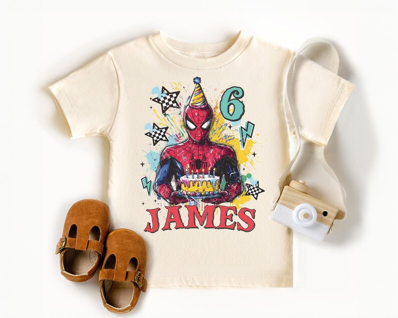 A white t-shirt with a Spiderman graphic and the number 6, along with brown baby shoes and a camera, all arranged on a white background.