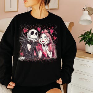 A woman wearing a black sweatshirt with a graphic design featuring a skeleton and a woman with a pink bow.