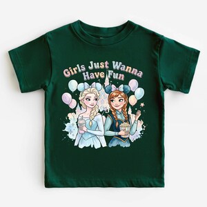 A green t-shirt with a graphic design featuring two cartoon girls, one blonde and one brunette, surrounded by balloons and a castle. The text above the girls reads "Girls Just Wanna Have Fun".