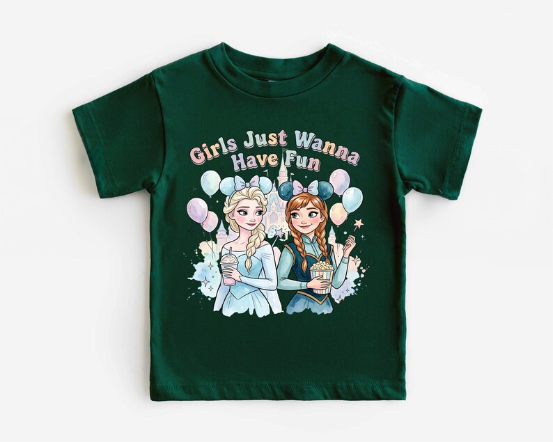 A green t-shirt with a graphic design featuring two cartoon girls, one blonde and one brunette, surrounded by balloons and a castle. The text above the girls reads "Girls Just Wanna Have Fun".