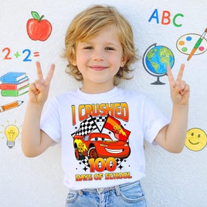 A young boy is smiling and making a peace sign with his hands, wearing a white t-shirt with a graphic of a red car and the text "I CRUSHED 100 DAYS OF SCHOOL".