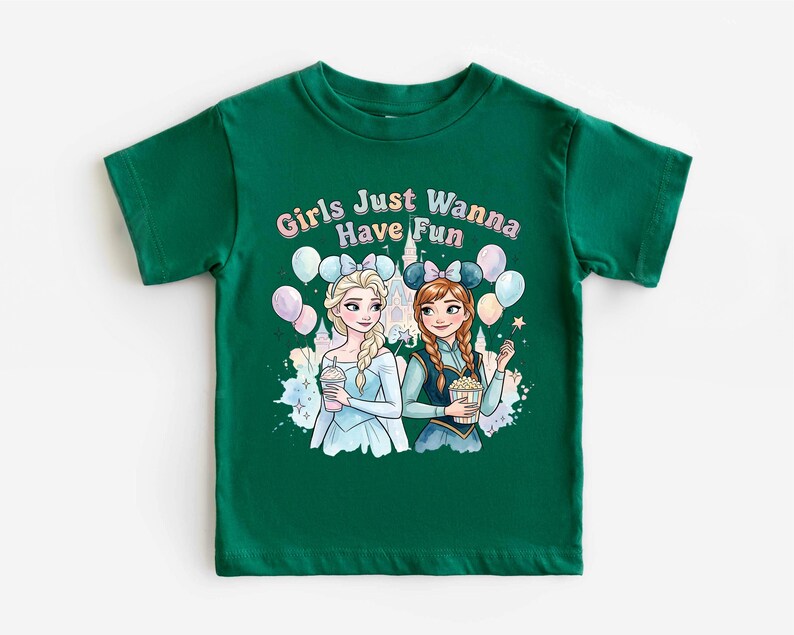 A green t-shirt with a graphic design featuring two cartoon girls, one blonde and one brunette, surrounded by balloons and a castle. The text above the girls reads "Girls Just Wanna Have Fun".