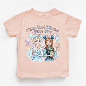 A pink t-shirt with a graphic design featuring two cartoon girls, one blonde and one brunette, surrounded by balloons and the text "Girls Just Wanna Have Fun".