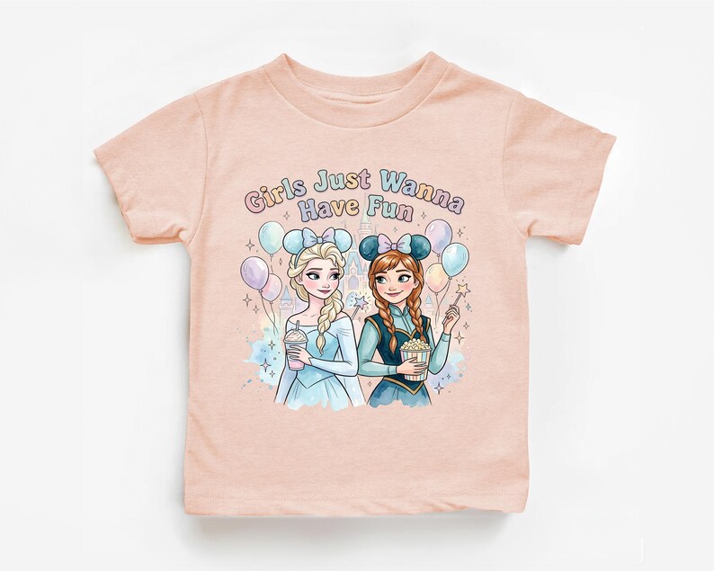A pink t-shirt with a graphic design featuring two cartoon girls, one blonde and one brunette, surrounded by balloons and the text "Girls Just Wanna Have Fun".