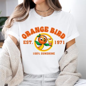 a woman sitting on a bed wearing an orange bird t - shirt
