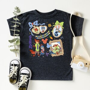 a black t-shirt with a colorful graphic design, including cartoon characters and various decorative elements. The t-shirt is placed on a white surface, accompanied by a pair of black and white sneakers, a white camera strap, and some greenery.