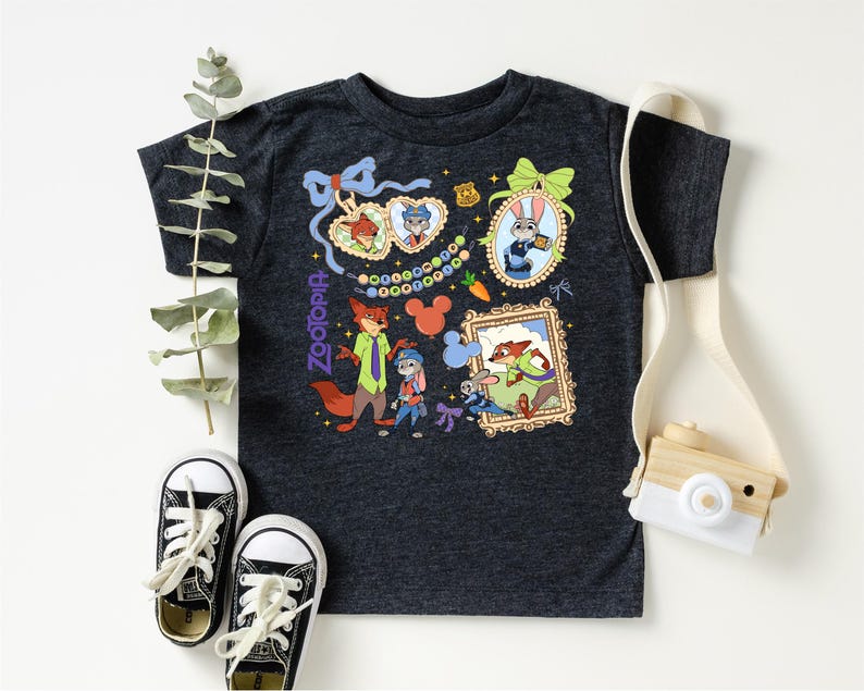 a black t-shirt with a colorful graphic design, including cartoon characters and various decorative elements. The t-shirt is placed on a white surface, accompanied by a pair of black and white sneakers, a white camera strap, and some greenery.