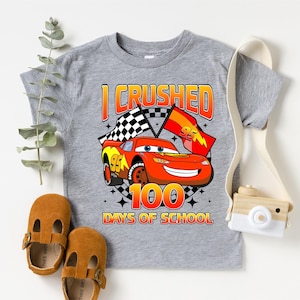 A gray t-shirt with a graphic of a red car and the text "I CRUSHED 100 DAYS OF SCHOOL" printed on it, along with a pair of brown sandals and a camera.