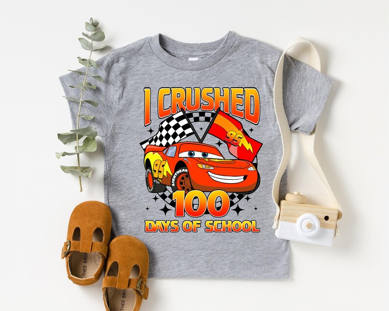 A gray t-shirt with a graphic of a red car and the text "I CRUSHED 100 DAYS OF SCHOOL" printed on it, along with a pair of brown sandals and a camera.