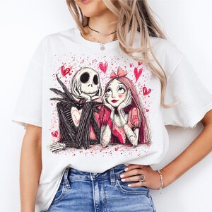 A woman wearing a white t-shirt with a graphic design featuring a skeleton and a woman with a heart-shaped bow on her head.