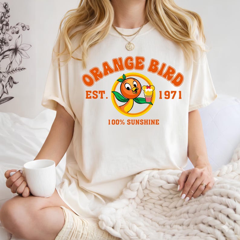 A woman wearing a white t-shirt with an orange bird graphic and text, sitting on a bed with a white blanket.