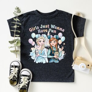 A black t-shirt with a graphic design featuring two cartoon girls, one blonde and one brunette, surrounded by balloons and the text "Girls Just Wanna Have Fun". The t-shirt is placed on a white surface, with a pair of sneakers, a camera strap, and some greenery visible in the background.