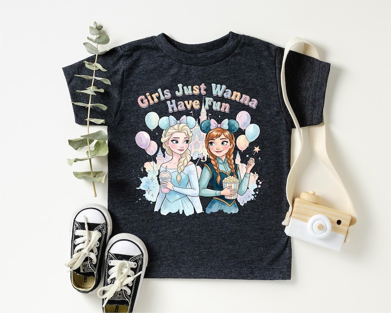 A black t-shirt with a graphic design featuring two cartoon girls, one blonde and one brunette, surrounded by balloons and the text "Girls Just Wanna Have Fun". The t-shirt is placed on a white surface, with a pair of sneakers, a camera strap, and some greenery visible in the background.