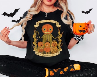 May include: Black t-shirt with a Halloween-themed graphic featuring a pumpkin-headed character and the text "WELCOME TO POTTSFIELD WHAT A WONDERFUL HARVEST." An orange pumpkin-shaped mug is held in the hand.