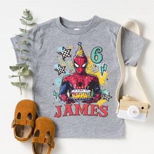 A gray t-shirt with a Spiderman graphic and the number 6, along with brown baby shoes and a camera, all arranged on a white background.
