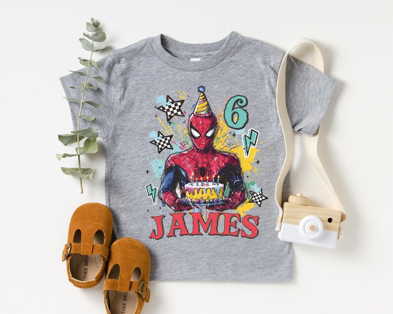 A gray t-shirt with a Spiderman graphic and the number 6, along with brown baby shoes and a camera, all arranged on a white background.