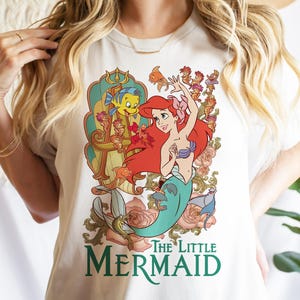 Op de afbeelding: Wit T-shirt met een kleurrijke afbeelding van Ariel uit De Kleine Zeemeermin. Het ontwerp toont Ariel met rood haar, een turquoise staart en een paarse schelpenbeha, samen met Botje en andere personages. De woorden "The Little Mermaid" zijn in het groen gedrukt.