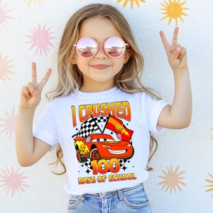 A young girl wearing a white t-shirt with a graphic of a red car and the text "I CRushed 100 DAYS OF SCHOOL" is making a peace sign with her hands.