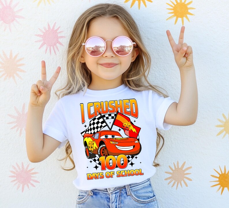 A young girl wearing a white t-shirt with a graphic of a red car and the text "I CRushed 100 DAYS OF SCHOOL" is making a peace sign with her hands.