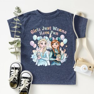 A blue t-shirt with a graphic design featuring two cartoon girls, one blonde and one brunette, surrounded by balloons and the text "Girls Just Wanna Have Fun". The t-shirt is placed on a white surface, with a pair of black and white sneakers, a white camera strap, and some greenery.
