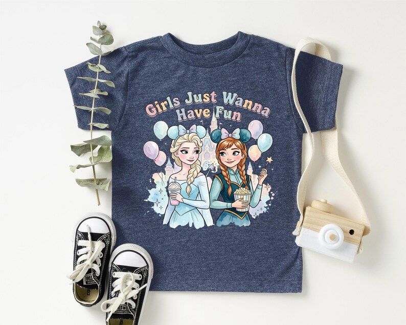 A blue t-shirt with a graphic design featuring two cartoon girls, one blonde and one brunette, surrounded by balloons and the text "Girls Just Wanna Have Fun". The t-shirt is placed on a white surface, with a pair of black and white sneakers, a white camera strap, and some greenery.