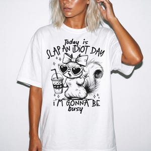 May include: A white t-shirt featuring a black graphic illustration of a squirrel wearing heart-shaped sunglasses and a bow, holding a drink. The text reads: Today is SLAP AN iDiOT DAY i'M GONNA BE busy. The design includes small star accents.