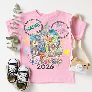 A pink Disney-themed t-shirt with a group of cartoon characters, including Mickey Mouse, Donald Duck, Goofy, and others, along with a pair of sneakers and a camera.